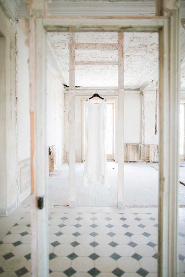 A getting-ready detail shot of a simple, column-style ivory wedding dress hanging on a black hanger in a doorframe within a derelict interior space. The room features heavily distressed white-painted walls with exposed plaster and brick, a vintage cast-iron radiator, and a classic black-and-white diamond-patterned tile floor. The composition is a wide shot taken through a doorframe, creating a layered, receding perspective with natural backlight from a window behind the dress. The overall styling aesthetic is minimal and editorial, contrasting the clean lines of the gown against the worn, textured architecture.