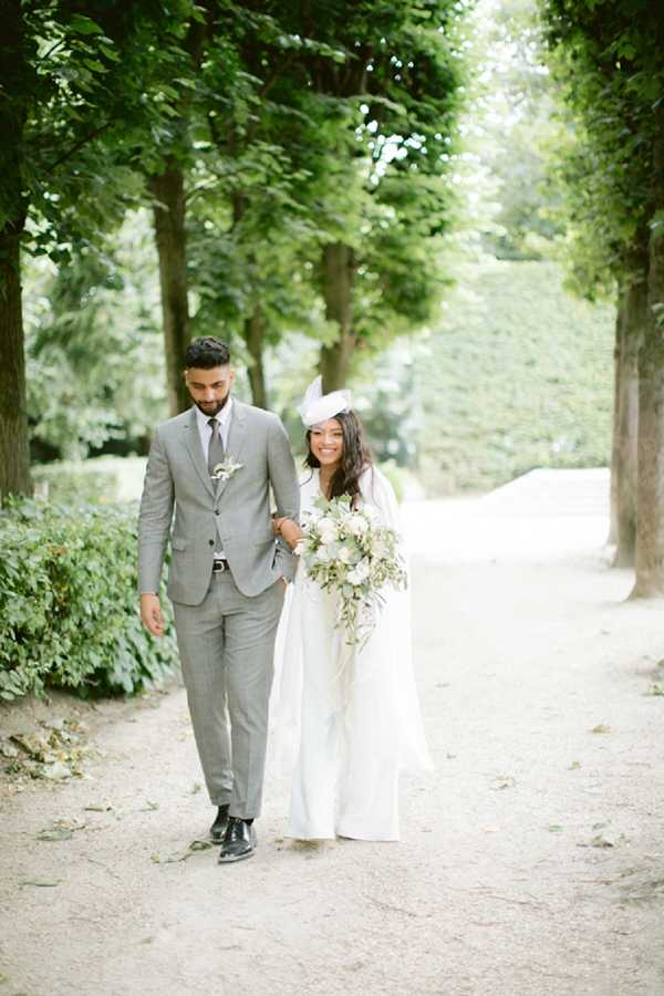 A couple walks arm-in-arm along a tree-lined gravel path in an outdoor formal garden setting, likely the grounds of a French chateau or park. The groom wears a light grey suit with a grey tie and a small white floral boutonniere, while the bride wears a long white gown and a white fascinator hat, carrying a loose, cascading bouquet of blush and ivory blooms with trailing greenery. The composition is a full-length portrait shot taken from a slight distance, with the manicured tree canopy creating a natural tunnel effect along the path. The overall styling is classic and refined with a soft, airy palette.