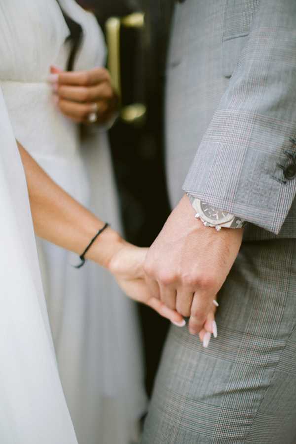 Close-up detail shot of a bride and groom holding hands. The bride is wearing a white dress with a flowing veil visible on the left, a thin black bracelet on her wrist, and has white-tipped nails; her other hand holds what appears to be a dark clutch with a gold accent. The groom is dressed in a light grey glen plaid suit and wears a silver chronograph watch. The composition is tightly cropped to focus on the hands and accessories, with the background softly blurred.