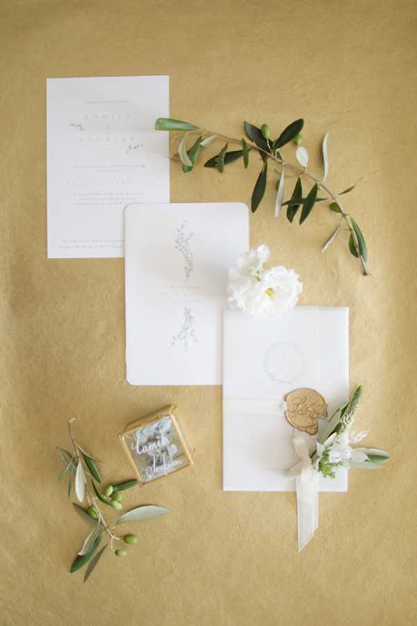 A flat lay detail shot of a wedding stationery suite arranged on a warm golden/ochre textured surface. The suite includes a white invitation card with minimal script typography, a smaller enclosure card featuring a delicate botanical illustration, and a white vellum envelope sealed with a round kraft wax seal bearing script lettering. Styling accents include a small acrylic or glass ring box with white script lettering, a white peony bloom, a white floral boutonniere with greenery and ribbon, and two olive branches with green olives scattered around the composition. The overall palette is white, ivory, and green against the golden background, with a classic and minimalist design aesthetic.
