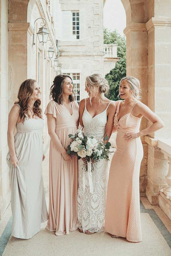 A bridal party portrait taken on an outdoor stone colonnade of a French chateau, featuring the bride and three bridesmaids laughing together. The bride wears a fitted ivory lace gown with spaghetti straps and an intricate embroidered pattern, holding a bouquet of white garden roses, ivory blooms, and eucalyptus greenery. The bridesmaids wear mismatched floor-length gowns in a coordinated soft palette: one in pale sage grey with a smocked bodice, one in blush pink with flutter sleeves and a wrap-style bodice, and one in peach with a textured fabric and knotted front detail. The shot is a full-length portrait with warm natural light and the chateau's limestone arched columns and windows visible in the background.