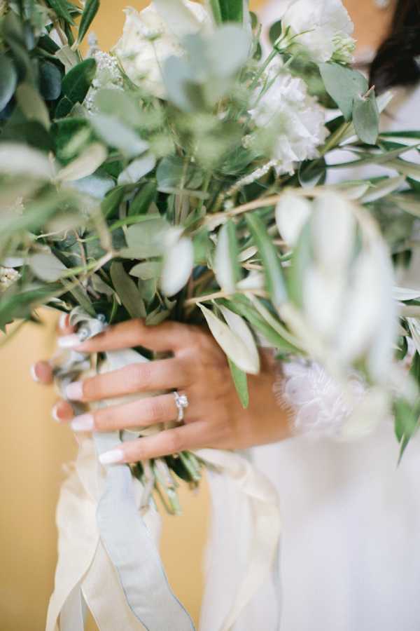 Close-up detail shot of a bride's hands holding a loosely arranged bouquet composed predominantly of olive branches, eucalyptus, and white flowers including lisianthus and small white filler blooms. The stems are wrapped with trailing ivory and pale sage silk ribbons. The bride wears a diamond solitaire engagement ring and a white garment with a feathered or fur-trimmed cuff visible at the wrist. Her nails are painted with a French manicure. The background is a warm ochre-yellow wall, softly out of focus.