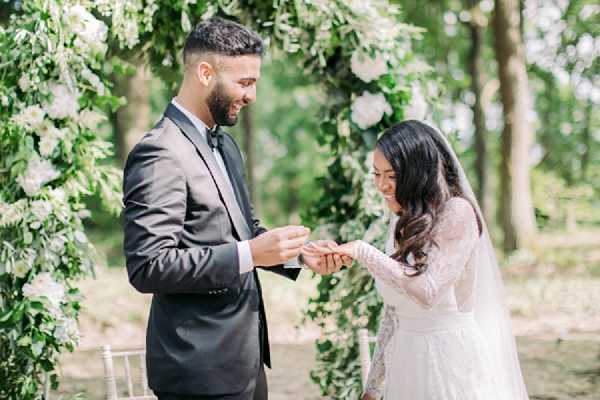 The groom is placing a ring on the bride's finger during an outdoor ceremony ring exchange. The setting is a wooded garden with trees visible in the background, and the couple stands in front of a large floral arch decorated with white blooms and lush green foliage. The groom wears a dark charcoal suit with a black bow tie, while the bride wears a long-sleeve lace white gown with a veil, her dark hair worn in loose waves. The styling is classic and refined, with an all-white and green floral palette. The image is a medium portrait shot with soft, bright natural lighting.