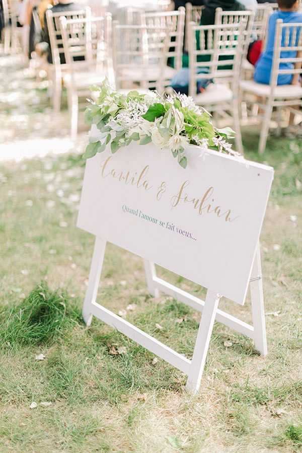 Close-up detail shot of an outdoor wedding ceremony welcome sign displayed on a white A-frame easel on a grass lawn. The white rectangular sign features the couple's names 'Camila & Julien' in gold calligraphy script, with the French phrase 'Quand l'amour se fait voeux...' printed below in smaller text. The top of the sign is decorated with a loose arrangement of green eucalyptus foliage and small white blooms. In the softly blurred background, rows of natural wood chiavari chairs are set up for the ceremony with a few guests already seated, suggesting the ceremony is about to begin. The overall styling is clean and modern with a white and gold color palette accented by greenery.