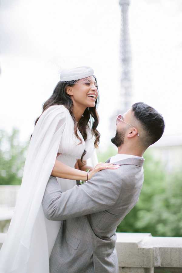 A couple portrait shot outdoors in Paris with the Eiffel Tower visible and softly out of focus in the background. The groom, wearing a light grey suit and tinted pink-lens sunglasses, lifts the bride while looking up at her; she laughs openly, looking upward. The bride wears a white bridal cape over what appears to be a fitted white or ivory outfit, paired with a small white pillbox-style hat with a birdcage veil — a modern, fashion-forward bridal look. The overall styling theme is chic and urban. Medium portrait composition with a shallow depth of field.