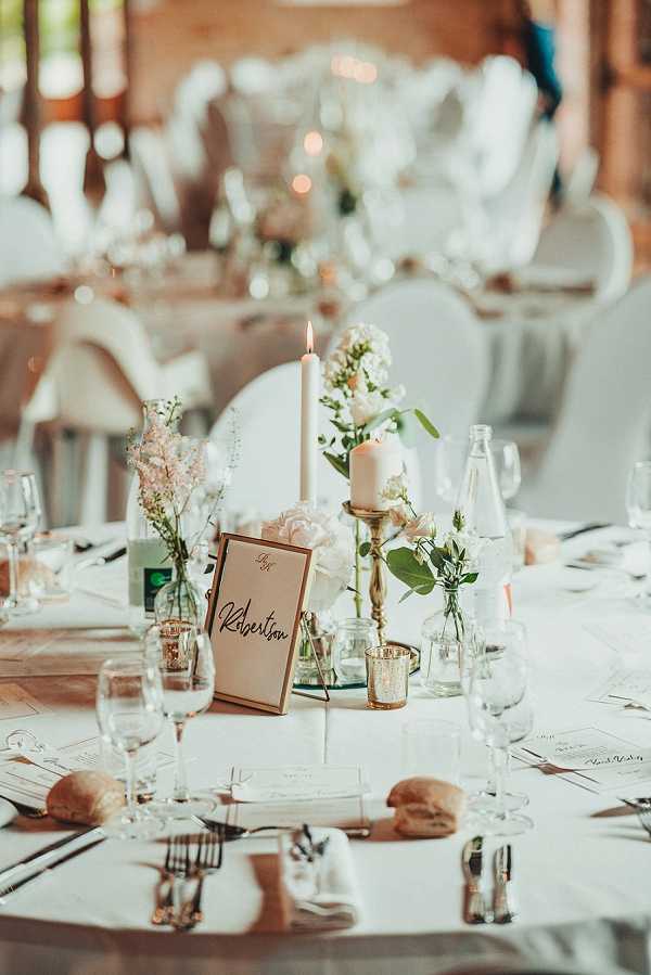A close-up detail shot of a wedding reception table setting inside what appears to be a barn or rustic indoor venue with warm wooden tones visible in the background. The round table is dressed in a white linen tablecloth and set with white folded napkins, silver cutlery, wine glasses, bread rolls, and printed menus. The centerpiece features a gold candlestick holder with a lit taper candle alongside a white pillar candle, small bud vases with blush pink roses, white lisianthus, pale pink astilbe, and greenery, plus gold tea light votives and a mirrored base. A framed table name card with gold border reads 'Robertson' in calligraphy script. In the softly blurred background, additional round tables with white ghost-style chairs are visible, decorated in a similar style, with warm ambient lighting creating a soft glow throughout the room. The overall decor palette is white, blush pink, and gold with a modern classic styling approach.