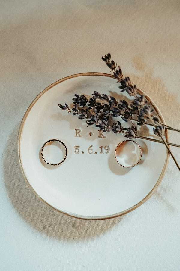 Close-up flat lay detail shot of two wedding rings resting on a small round white ceramic ring dish with a gold rim, stamped with the initials 'R + K' and the wedding date '5.6.19'. One ring features a delicate milgrain or diamond-set band, while the other is a plain gold band. A small sprig of dried lavender is laid across the dish. The dish is placed on a cream linen surface, giving the composition a soft, muted tone. The overall styling is understated and romantic, consistent with a rustic or natural aesthetic.