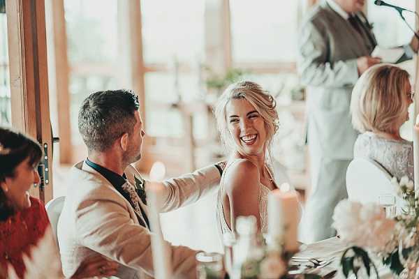 The bride and groom are seated at the head table during wedding speeches, with the bride laughing and turning toward the groom, who has his arm around her shoulder. The bride wears a low-back white gown with her blonde hair loosely pinned up, while the groom is dressed in a light beige suit with a floral boutonniere. The setting is an indoor reception room with large windows letting in warm natural light, and white floral centerpieces and candles are visible on the table in the foreground. A speaker holding a microphone stands partially visible in the background alongside other seated guests, suggesting a relaxed, candid atmosphere. The image is a mid-range candid portrait shot with warm, golden-toned lighting.