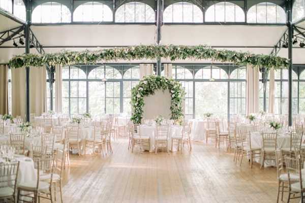 Wide shot of an indoor wedding reception hall set up for a seated dinner, with no guests present. The venue features large arched steel-framed windows along the walls and ceiling that flood the space with natural light, and a pale hardwood floor. Round tables are dressed in white linen tablecloths and surrounded by gold chiavari chairs, with low white floral centerpieces. A large circular floral arch made of lush greenery and soft white and blush blooms serves as a focal backdrop against a white wall. Overhead, a suspended horizontal garland of trailing greenery runs across the iron ceiling structure. The overall decor palette is white, ivory, and green with gold accents, in a classic garden-inspired style. Potential venue feature image.