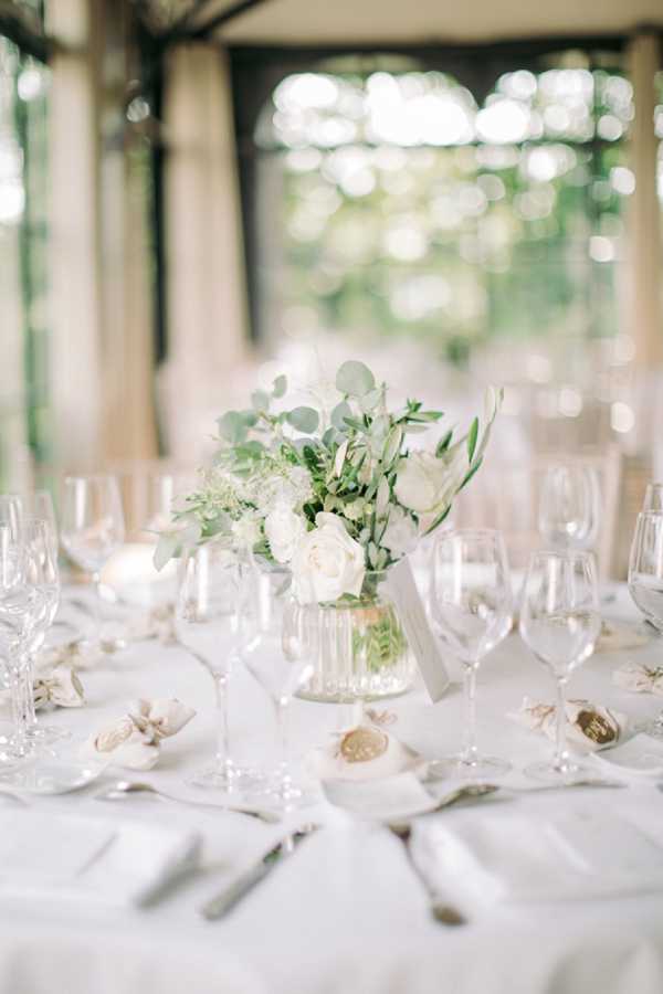Close-up detail shot of a wedding reception table setting in what appears to be a covered outdoor pavilion or veranda with tall columns and draping curtains visible in the background. The round table is dressed in a white linen tablecloth with white folded napkins, silver cutlery, and multiple clear crystal wine and water glasses per place setting. The centerpiece is a low, ribbed glass vase holding white roses, white ranunculus, eucalyptus, and olive branch greenery. Small gold wax seal place card holders are visible at each setting, and a small white table number card is tucked into the centerpiece arrangement. The overall decor palette is white and gold with greenery accents, reflecting a classic, clean aesthetic.