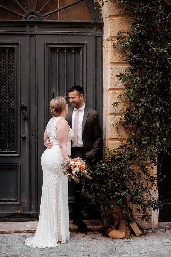 A couple portrait taken outdoors on a cobblestone street in front of a large dark charcoal-grey double door with ornate ironwork and a fanlight above. The bride wears a fitted, lace or beaded white gown with a short train and has her blonde hair pinned up with a small decorative hair accessory; she holds a bouquet of autumnal flowers in terracotta, rust, peach, and dusty orange tones with greenery and eucalyptus. The groom wears a dark brown suit with a light blush-pink tie and has his arm around the bride's waist, smiling down at her. The setting has a classic Parisian or southern French urban character, with ivy climbing the warm sandstone wall beside the doorway. Medium portrait shot, slightly from behind the bride, capturing both figures at full length.