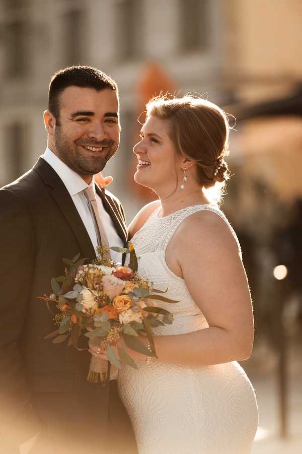 A couple portrait taken outdoors in warm golden-hour light, with a blurred architectural background. The bride wears a fitted ivory beaded halter-neck gown with a racerback style and pearl drop earrings, with her hair in a low updo. She holds a loose, garden-style bouquet featuring peach and burnt orange roses, blush blooms, dusty blue accents, and eucalyptus greenery. The groom wears a charcoal suit with a white dress shirt and a light grey tie. The two are facing each other and smiling, with the groom looking toward the camera. Close-up couple portrait shot with shallow depth of field.