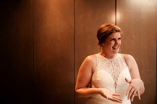 A close-up portrait of a bride laughing and looking to one side, standing against a large dark bronze or copper-toned panel wall. She is wearing a sleeveless, high-neck wedding dress featuring an intricate beaded and embroidered geometric pattern across the bodice. Her hair is styled up, and she wears drop earrings and a delicate bracelet. The warm ambient lighting casts a golden tone across the scene, highlighting her expression and the texture of the beadwork on her dress.