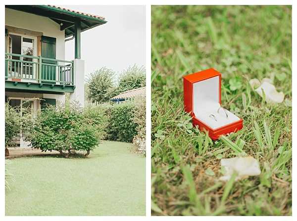 A two-panel image combining a venue exterior shot and a ring detail close-up. The left panel shows a white two-story building with dark green balcony railings and shutters, surrounded by a manicured lawn and dense shrubs, photographed as a wide exterior shot. The right panel is a close-up detail shot of two plain gold or white metal wedding bands displayed in an open orange ring box with a white interior, placed on grass with a few white flower petals scattered nearby. The overall styling is simple and understated with no elaborate floral or decor elements visible.