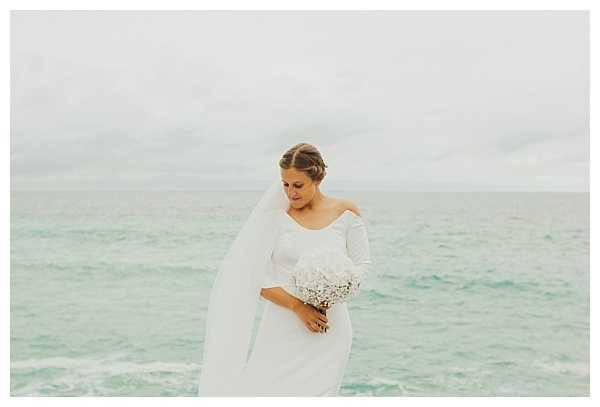 A bridal portrait taken outdoors with the ocean as a backdrop, shot on an overcast day with soft, muted light. The bride wears a fitted white long-sleeve gown with an off-the-shoulder neckline and a long cathedral-length veil draped over one shoulder, her hair styled in an updo. She holds a compact bouquet of white flowers, likely hydrangeas and baby's breath, and looks downward with a calm expression. The composition is a medium portrait shot with the pale turquoise-green sea filling the background.