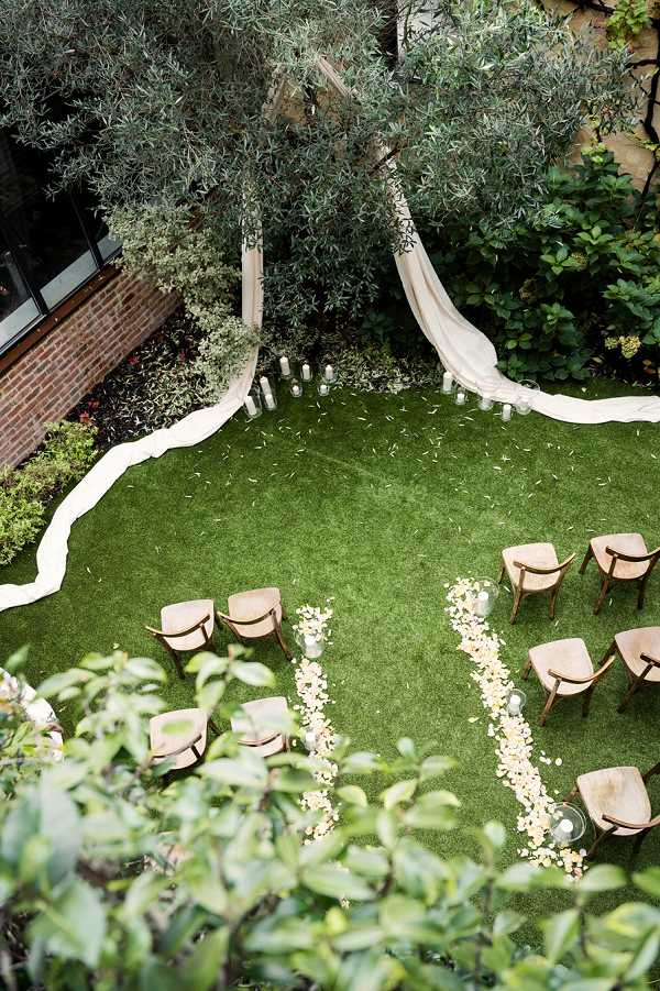 An aerial shot of an outdoor wedding ceremony setup in a compact courtyard garden with a manicured lawn. The ceremony space features wooden chairs with curved backs arranged in two short rows on either side of a central aisle, which is lined with scattered cream and pale yellow flower petals and small glass globe vases. At the altar end, two lengths of flowing ivory fabric are draped from an olive tree, creating a natural canopy effect, flanked by white pillar candles of varying heights placed along the ground. The overall decor palette is ivory, cream, and natural wood, giving the setup a clean, minimalist feel with organic elements.