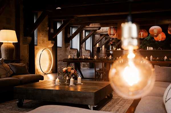 An indoor wedding lounge or cocktail hour setup inside a rustic venue with exposed dark wooden beams and stone walls. The space features a dark wood coffee table styled with pillar candles and a small arrangement of peach and coral flowers, surrounded by low upholstered sofas in neutral linen and dark brown velvet. Warm ambient lighting comes from a large Edison-style globe bulb in the foreground, cluster pendant lights with warm orange globe shades hanging from the ceiling, a floor lamp with a woven shade, and rows of small candles arranged on a shelf or bar area in the background. The decor palette is warm and moody, combining earth tones, burnt orange, and candlelight, with a rustic-industrial aesthetic. Wide interior shot with a blurred foreground bulb creating depth. Potential venue feature image.