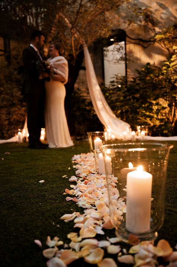 A couple shares a close embrace at the altar during or just after an outdoor evening ceremony, set in a garden with mature trees and greenery. The aisle is lined with scattered blush and white rose petals alongside glass hurricane candle holders containing large white pillar candles, creating warm ambient lighting throughout the space. The bride wears a fitted white gown with a white fur or feather wrap over her shoulders and carries a small bouquet, while the groom is dressed in a dark suit; a black-framed arch draped with sheer ivory fabric is visible behind them. The composition is a depth-of-field shot taken from ground level, with the candles and rose petals sharp in the foreground and the couple softly blurred in the background, emphasizing the candlelit decor in a classic, romantic evening ceremony style.