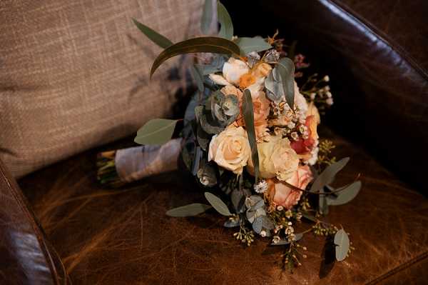 Close-up detail shot of a bridal bouquet resting on a worn brown leather armchair with a textured linen cushion visible in the background. The bouquet features peach and blush roses, small terracotta-toned blooms, white wax flower, eucalyptus leaves, and trailing greenery in a loose, organic arrangement. The stems are wrapped in what appears to be a dusty grey-blue ribbon or fabric. The overall color palette is warm and earthy, consistent with a rustic or boho styling theme.