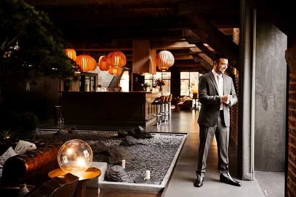 A groom in a charcoal grey suit with a white dress shirt and tie stands near a doorway or large open panel inside a contemporary venue, smiling and holding what appears to be a small glass. The interior space features exposed dark wood ceiling beams, warm amber and orange paper lantern pendant lights hanging above a bar area with leather bar stools, and a decorative indoor rock garden or gravel feature running along the floor. A large glass globe lamp and small candles sit on a low wooden side table beside a leather sofa, contributing to the moody, warm ambient lighting. The overall decor aesthetic is modern with Asian-influenced design elements, shot as a wide environmental portrait.