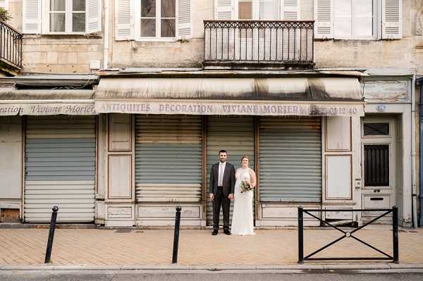 A bride and groom stand together on a sidewalk in front of a closed French antique and decoration shop called 'Antiquités Décoration Viviane Morier,' with shuttered storefronts and a weathered facade visible behind them. The groom wears a dark charcoal suit with a tie, and the bride wears a slim, ivory gown and holds a small bouquet with greenery and what appear to be neutral-toned blooms. The couple is positioned centrally in a wide environmental portrait shot that deliberately frames them against the peeling paint, rolled awnings, and aged urban streetscape of a French town. The styling is understated and the urban location gives the portrait a candid, editorial feel.