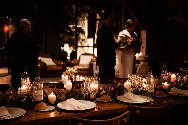 A candlelit wedding reception dinner table is shown in the foreground, set with white plates, linen napkins, wine glasses, and wine bottles, with a low greenery runner and clusters of white pillar candles creating warm amber lighting across the wooden table. In the softly blurred background, two figures — one in dark clothing and one in white, likely the bride holding a bouquet with muted floral tones — stand near a warmly lit interior space furnished with lounge seating. The overall decor palette is dark and moody with warm candlelight as the primary light source, giving the setting an intimate, rustic-romantic atmosphere. This is a wide reception detail shot with a shallow depth of field emphasizing the table setup in the foreground.