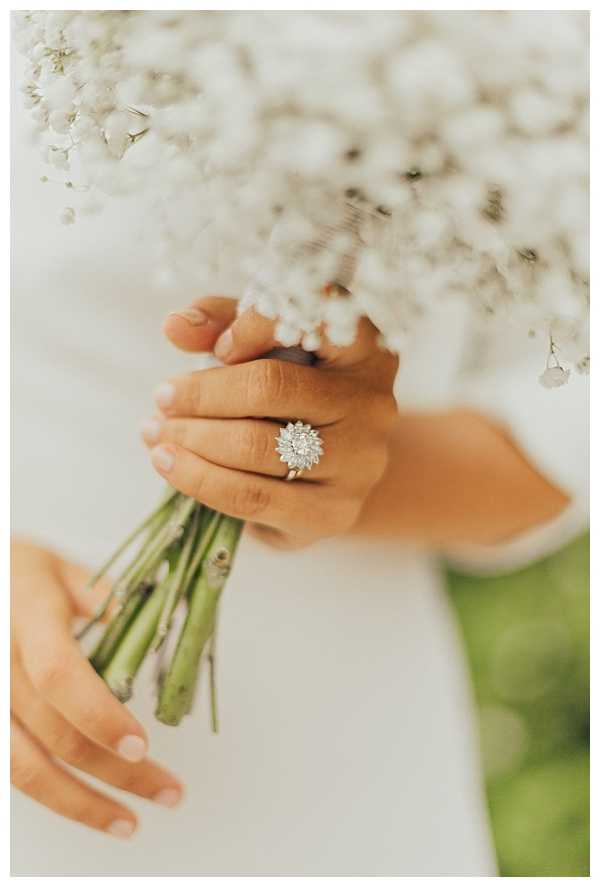 Close-up detail shot of a bride's hands holding a all-white baby's breath bouquet with green stems, wearing a statement diamond cluster engagement ring with a starburst or floral setting in a silver-toned metal. The bride is dressed in a white gown, visible at the edges of the frame. The bouquet fills the upper portion of the image, slightly out of focus, drawing attention to the ring as the focal point. The overall styling is minimal and monochromatic, with the all-white baby's breath bouquet complementing the simple bridal look.