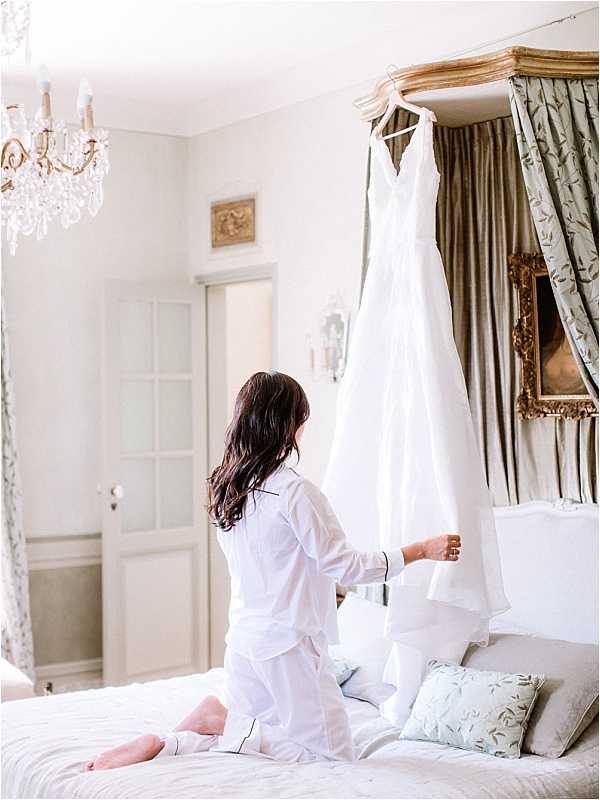 A bride is kneeling on a bed in a getting-ready scene, reaching toward her wedding dress, which hangs from a gilded four-poster canopy bed frame. She is wearing white pajamas with black piping trim and has long dark hair. The wedding gown is an ivory sleeveless A-line style with a V-neckline. The room is decorated in a classic French chateau style, featuring a crystal chandelier, ornate gold-framed mirror, sage green floral canopy drapes, light blue and sage embroidered throw pillows, and white-painted wood-paneled doors with gilt wall sconces. The overall decor palette is soft ivory, sage, and gold. Medium-distance portrait shot from behind the bride.