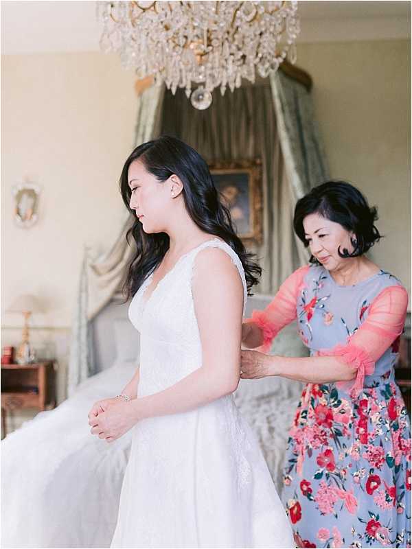 A getting-ready scene captured indoors in what appears to be a chateau bedroom, where an older woman in a blue and red floral dress with coral sheer sleeves fastens the back of the bride's white lace gown. The bride, with long dark hair worn loosely to one side, stands with hands clasped in front of her wearing a sleeveless V-neck white lace dress. The room features a crystal chandelier overhead, a canopied bed with sage green draping in the background, antique furnishings, and framed artwork on the walls, suggesting a classic French chateau setting. The portrait-style composition is shot from a slight side angle, with soft, warm natural light filling the room.
