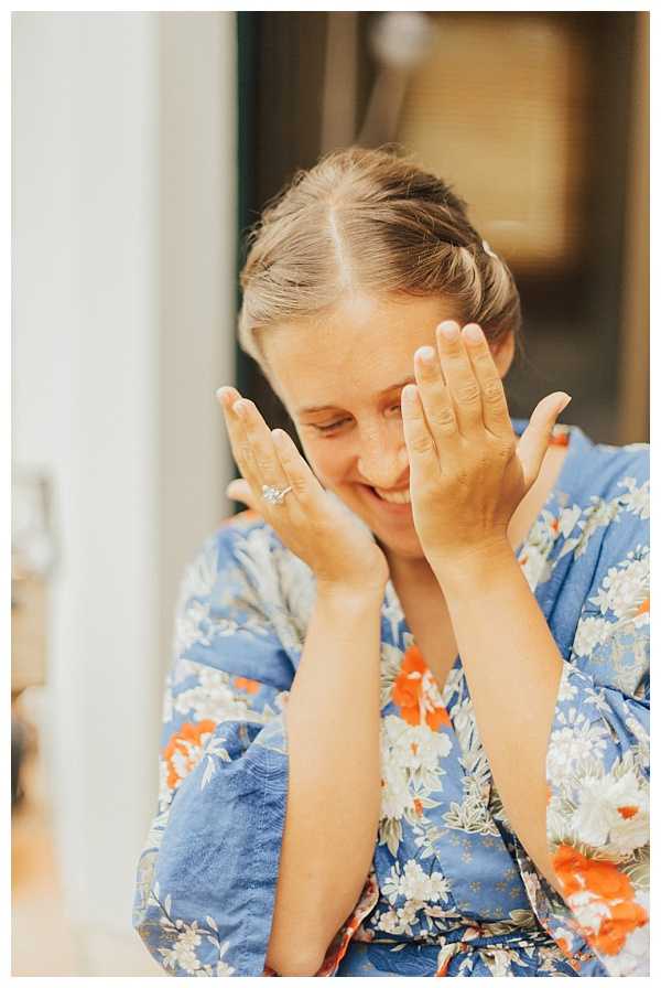 A getting-ready moment captured indoors, showing the bride laughing with her hands raised near her face, appearing to react to an emotional or joyful moment. She is wearing a blue floral robe with orange and white flower print, and a diamond engagement ring is visible on her left hand. Her hair is partially pinned up. The shot is a close-up portrait with a soft, warm background that is blurred out of focus.