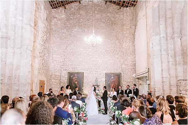 A wedding ceremony taking place inside a historic stone chapel or converted church with exposed limestone walls and a high vaulted ceiling. The couple stands at the altar facing each other, flanked by a bridal party of approximately six people in black formal attire, with two large religious oil paintings hanging on the stone wall behind them and a small crystal chandelier overhead. Approximately 60–80 seated guests fill wooden chairs arranged in rows along a central aisle, which is lined with floral arrangements featuring pink, white, and purple blooms. The ceremony space is styled with a classic, romantic aesthetic, and the wide-angle shot captures the full interior from slightly elevated behind the guests, giving a clear view of the entire ceremony setup.