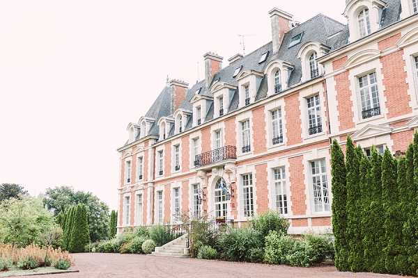 Wide exterior shot of a large French chateau featuring red brick facade with white stone trim, mansard roof with dormer windows, and ornate wrought-iron balconies. The building is three stories tall with symmetrical window arrangements and a central entrance framed by decorative stonework and steps. The foreground shows a gravel courtyard with manicured hedging and ornamental plantings. Potential venue feature image.