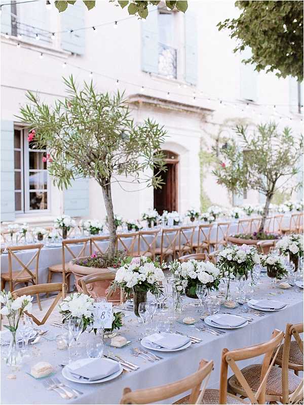 An outdoor wedding reception setup in a courtyard in front of a French manor house with pale blue shutters and warm stone facade. Multiple long banquet tables are dressed in dusty blue-grey linen and set with white ceramic plates, silver flatware, and crystal glassware, with natural wood cross-back chairs. Centerpieces consist of low, lush arrangements of white roses, white ranunculus, and white peonies in dark vessels, interspersed with single-stem bud vases and small candles. Mature olive trees planted in the courtyard are incorporated into the table layout, and string lights are strung overhead. The overall decor palette is soft blue-grey, white, and warm natural wood, giving a classic Provençal feel. Wide establishing shot showcasing both the table details in the foreground and the full reception layout extending toward the venue. Potential venue feature image.