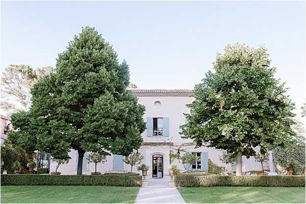 Wide exterior shot of a French country manor or bastide, featuring a two-story pale stone facade with dusty blue-grey shutters and a terracotta tile roof. A straight pathway leads to the arched front entrance, flanked symmetrically by two large mature trees and trimmed hedgerows. No people are visible; the shot is purely architectural, taken during soft early evening light. Potential venue feature image.