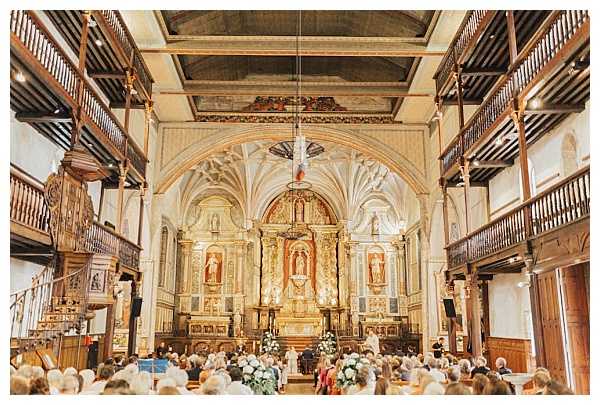 A wedding ceremony is taking place inside a historic French church featuring an ornate gilded altar, vaulted ceiling with fan tracery, and wooden upper gallery balconies running along both sides of the nave. The couple and officiant stand at the altar, surrounded by white and green floral arrangements, while a large congregation of approximately 80–100 guests fills the wooden pews. The interior architecture includes gold baroque altarpiece detailing, painted ceiling panels, and carved wooden furnishings. The shot is taken from the rear of the nave as a wide-angle view looking toward the altar, capturing the full length of the ceremony space. Potential venue feature image.