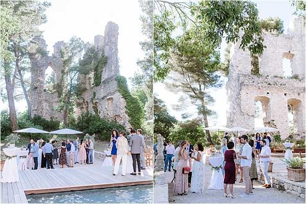 A cocktail hour taking place outdoors at the ruins of a historic stone castle or château, shown in two wide-shot frames. Approximately 30–40 guests are mingling on and around a wooden deck platform positioned beside a reflective pool or water feature, with the crumbling stone tower ruins as a dramatic backdrop. Guests are dressed in smart-casual summer attire including a white bridal gown, a blue dress, a burgundy dress, and light linen suits. White market umbrellas provide shade over the deck area, and potted plants are visible near the ruins. The overall styling is relaxed and classic, taking full advantage of the historic architectural setting. Potential venue feature image.
