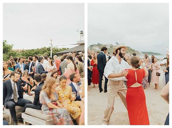 A diptych of two wide shots capturing a cocktail hour or reception on a coastal outdoor setting. The left image shows a large crowd of approximately 40-50 guests mingling and seated on lounge furniture on what appears to be a terrace or beach-adjacent deck, with guests dressed in smart casual and formal attire including navy suits, floral dresses, and colorful outfits. The right image shows a man in a white linen shirt, khaki trousers, and a straw hat dancing with a woman in a red open-back midi dress on a sandy beach, with additional guests visible in the background. The overall styling is relaxed and summery with a coastal, informal atmosphere, and the backdrop shows a rocky coastal headland and sandy beach.