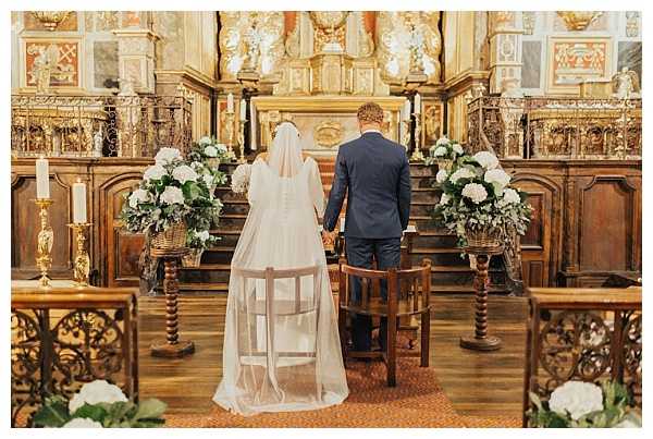 A bride and groom stand hand-in-hand at the altar during a religious ceremony inside an ornate historic chapel with gilded baroque woodwork and carved altarpiece. The bride wears a white gown with a long cathedral-length veil, and the groom is dressed in a navy blue suit. Two large floral arrangements in wicker baskets flank the altar steps, composed of white hydrangeas and green foliage. The shot is taken from behind the couple in a wide, symmetrical composition, with wooden pews and wrought-iron kneelers visible in the foreground.