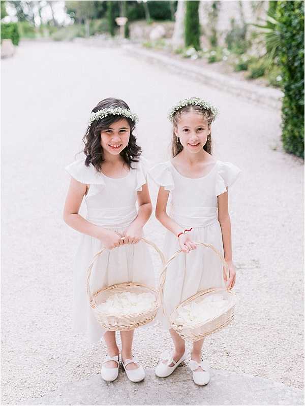 Two flower girls stand together on a gravel driveway lined with cypress trees and garden greenery, posing for a portrait. Both girls wear white flutter-sleeve dresses with white Mary Jane shoes and floral crowns made of baby's breath, and each holds a wicker basket filled with white flower petals. The setting appears to be the grounds of a French chateau or villa, with a classic, garden-style aesthetic. The image is a mid-range portrait shot with a soft, light-toned finish.