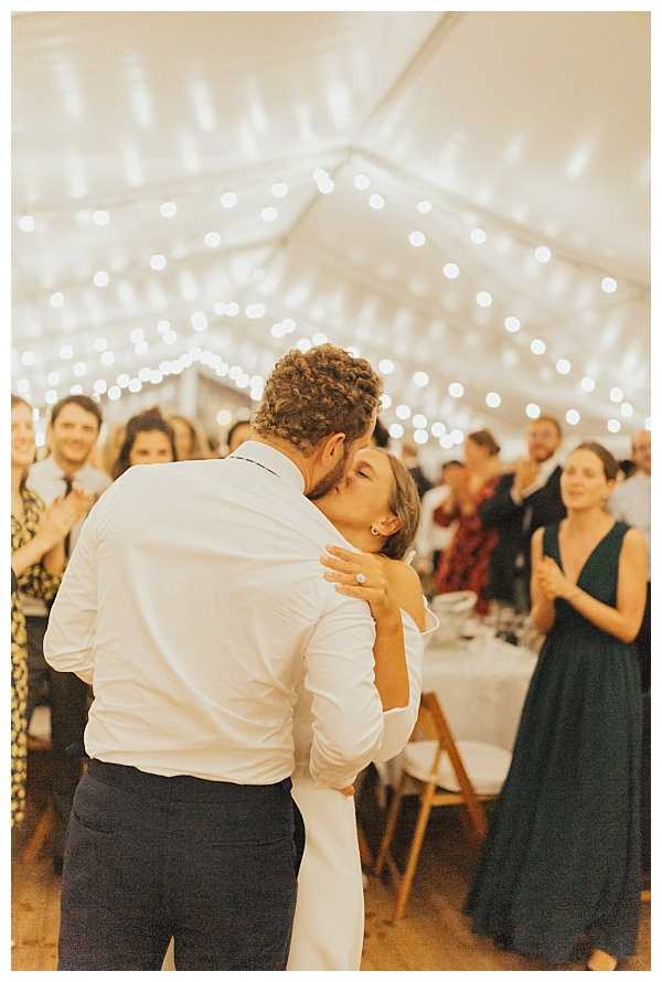 The bride and groom share a kiss on the dance floor during what appears to be their first dance at an indoor reception held under a white marquee tent strung with warm fairy lights across the ceiling. The groom wears a white dress shirt and dark trousers, while the bride wears a long-sleeved white gown and is visible wearing a diamond ring. Approximately 15–20 guests are visible in the background, clapping and watching, with one woman in a deep teal floor-length dress standing prominently to the right. The shot is a close portrait taken from behind the groom, with the background intentionally blurred, drawing focus to the couple's embrace.