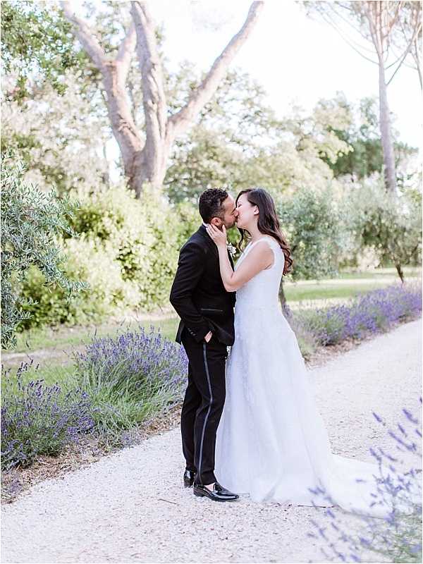 A couple shares a kiss during outdoor wedding portraits on a gravel path lined with blooming lavender bushes. The bride wears a sleeveless, fitted white gown with a short train and has long, wavy dark hair worn down; the groom wears a black tuxedo with satin side-stripe trousers and a small white boutonniere on his lapel. The setting appears to be a Provençal garden or estate grounds, with olive trees and tall pines visible in the soft, bright background. The shot is a full-length portrait with a classic, light-toned film-style finish.