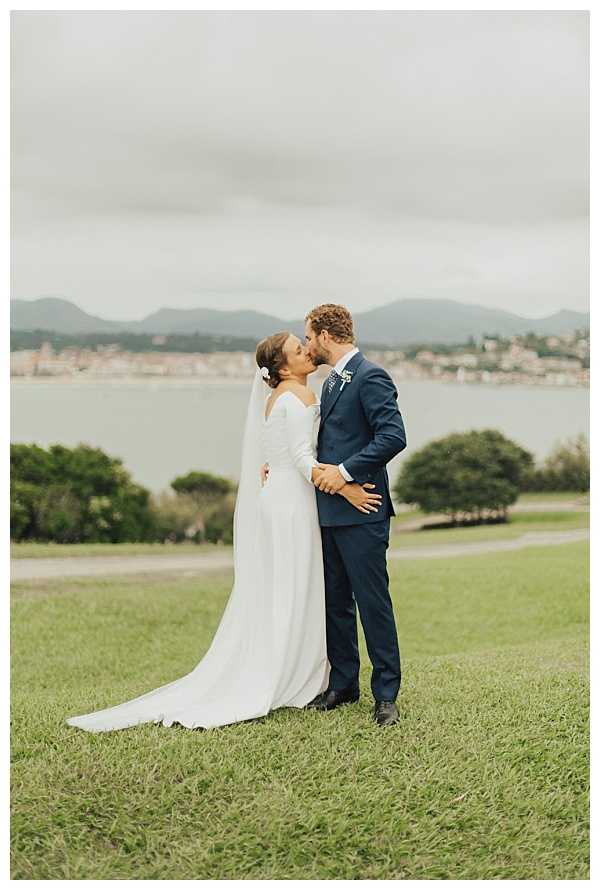 A couple sharing a kiss during outdoor wedding portraits on a grassy hillside with a coastal bay and distant mountains visible in the background. The bride wears a long-sleeved, fitted ivory crepe gown with a cathedral-length train and a small white floral hair accessory, paired with a sheer veil. The groom wears a navy blue suit with a small boutonniere. The shot is a full-length portrait taken at mid-distance, capturing both figures and the scenic waterfront backdrop behind them.