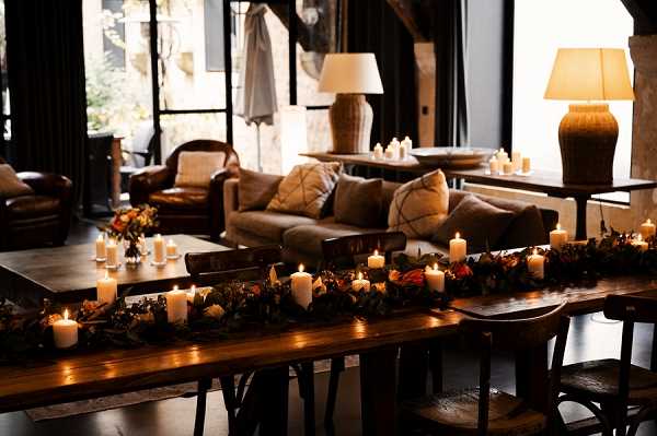 Detail shot of a wedding reception or cocktail lounge area styled in a warm, rustic aesthetic. A long dark wood trestle table in the foreground is decorated with a dense garland runner of dark foliage, deep red and burnt orange florals, and multiple lit ivory pillar candles of varying heights. In the background, a lounge seating area features brown leather armchairs and a neutral-toned sofa with textured cushions, with additional candles and small floral arrangements on low tables. Two large table lamps with woven bases and warm-toned shades provide ambient lighting alongside the candlelight, creating a moody, low-lit interior atmosphere. The overall decor palette is deep and autumnal — dark greens, burgundy, rust, and warm amber tones — consistent with a rustic or organic wedding styling theme.