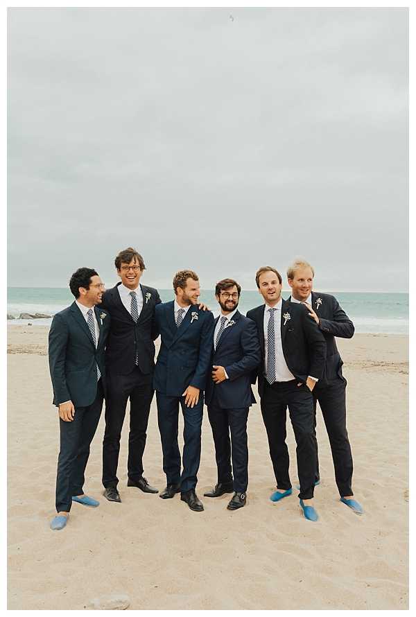 A group portrait of the groom and five groomsmen standing together on a sandy beach, posing casually and laughing. All six men wear navy or dark suits with patterned ties in shades of blue and grey, and small white floral boutonnieres; notably, two of the men on the right are wearing light blue espadrilles instead of dress shoes. The setting is an overcast beach with the ocean visible in the background. The shot is a full-length wide portrait with a relaxed, candid feel.