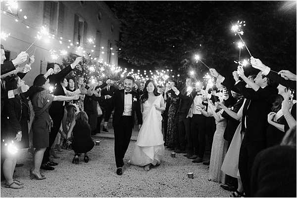 A black-and-white image of a couple making their grand exit at night, walking through a tunnel of lit sparklers held by two rows of guests. The bride wears an off-the-shoulder ball gown and the groom is dressed in a classic black tuxedo with bow tie; both are smiling and walking hand in hand. Approximately 30–40 guests line the path on a gravel driveway in front of what appears to be a chateau or manor building visible in the background. The sparklers create bright, high-contrast light trails against the dark night sky, giving the image strong tonal contrast typical of nighttime B&W photography. Wide shot with the couple centered in the frame.