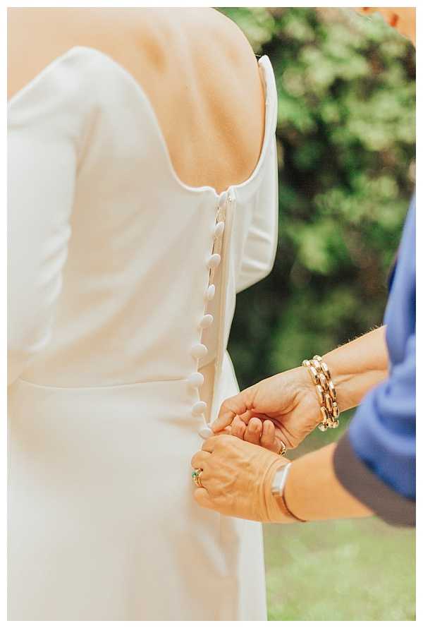A getting-ready moment captured in a close-up portrait shot outdoors, showing the back of a bride's ivory long-sleeve wedding dress with a row of fabric-covered buttons running down the spine. A second person, wearing a periwinkle blue outfit and gold chain bracelet with a green gemstone ring, is fastening the buttons at the lower back. The composition is tightly framed on the hands and dress detail, with a softly blurred green foliage background.