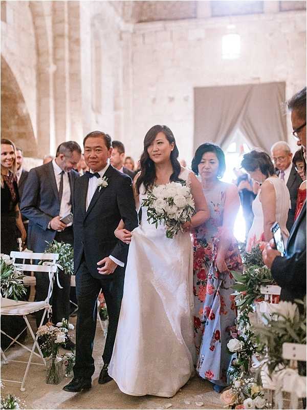 The bride is walking down the aisle during an indoor ceremony, escorted on each side by two family members — a man in a black tuxedo with a white boutonniere on her left and a woman in a light blue floral-print dress on her right. The bride wears a sleek, ivory satin gown with a structured silhouette and carries a round bouquet of white peonies and garden roses with trailing greenery. The ceremony is held inside a historic stone chapel or abbey, with tall stone walls and arched architecture visible in the background. The aisle is lined with white folding chairs and low floral arrangements in small vases featuring white blooms and green foliage. Guests stand on both sides watching the processional, with warm ambient lighting illuminating the space. The shot is a medium wide portrait taken from ground level facing the entrance.