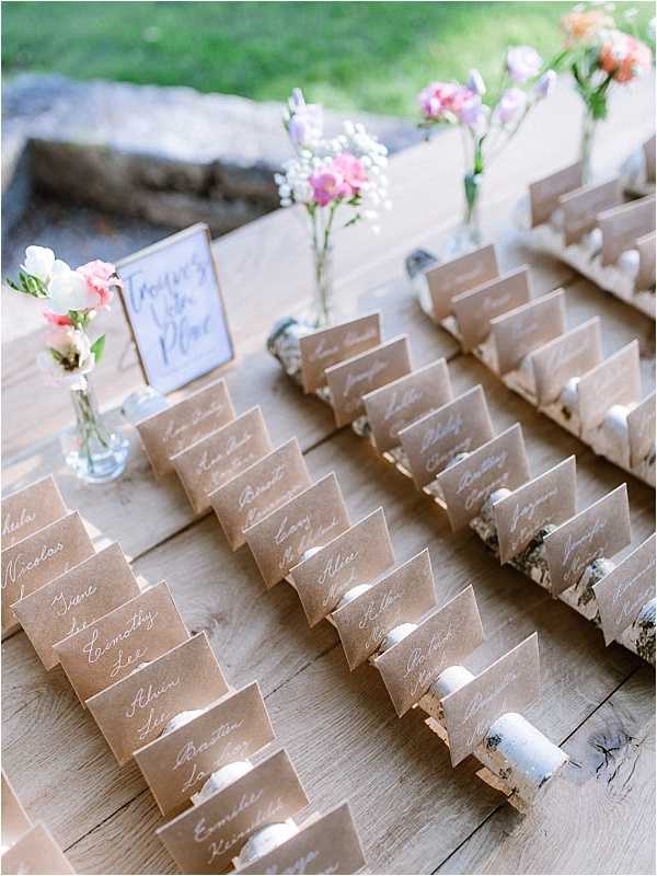 Close-up detail shot of a wedding seating chart escort card display arranged on a rustic wooden surface outdoors. Kraft paper cards with white calligraphy script are held upright by small birch wood log slice holders, arranged in neat rows across the table. A small handwritten sign reading 'Trouver Votre Place' (Find Your Seat) is propped among the cards, accompanied by small bud vases containing pink, coral, lavender, and white wildflower-style blooms including sweet peas and baby's breath. The overall styling is rustic-boho with a natural, earthy palette of kraft brown, white, and soft pastel florals.