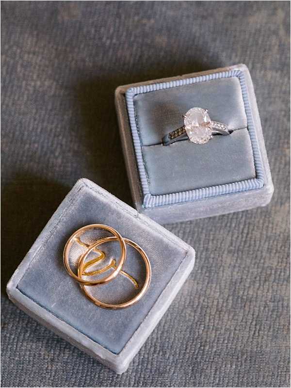 Close-up flat lay detail shot of two dusty blue velvet ring boxes displayed side by side on a muted grey textile surface. The top box holds a solitaire engagement ring featuring an oval-cut diamond on a white gold band with pavé-set stones along the shoulders. The bottom box contains two rose gold wedding bands, one of which appears to have an open, cutout initial or monogram design. The soft blue velvet of the boxes provides a cohesive color palette tying both pieces together.