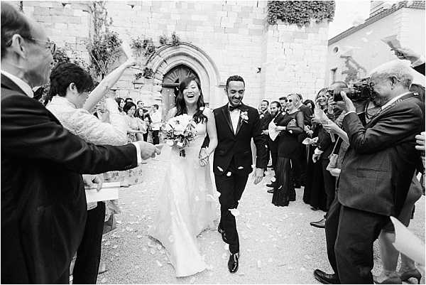 Black-and-white wide shot of a bride and groom exiting a stone chapel after their ceremony, walking through a confetti or petal send-off tunnel formed by approximately 20-25 guests on either side. The bride is laughing and wearing a full-length ballgown with a flowing skirt, carrying a round bouquet, while the groom wears a dark tuxedo with a bow tie and boutonniere. The chapel entrance features a rounded Romanesque stone archway in the background, suggesting a historic French church or chateau chapel. The image has strong contrast with bright highlights and deep shadows, and captures a candid, joyful moment mid-stride.